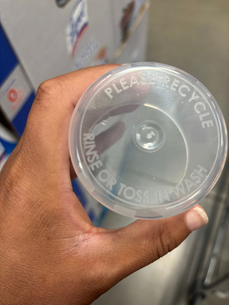 A hand holding a clear laundry detergent cap, which says on the top “please recycle” and on the bottom “rinse or toss in wash”