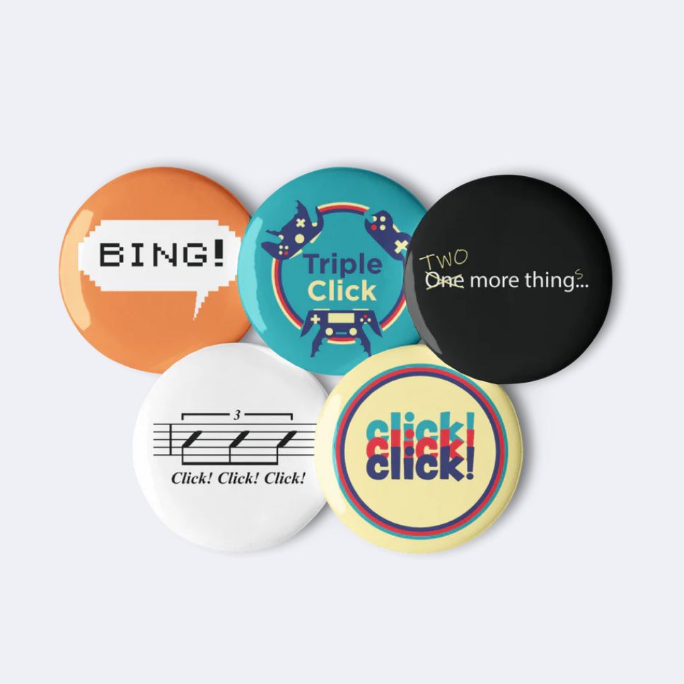 Set of five round pins.
#1: Orange pin with pixelated word bubble and black text reading BING!
#2: Triple Click logo
#3: Small white sans serif text reading "One more thing..." The text has been corrected in what looks like yellow pencil to read "TWO more things..."
#4: Musical notation for a percussion triplet, with italic text below reading "Click! Click! Click!"
#5: Vertically overlapping text in teal, red, and dark blue reading "click! click! click!"
