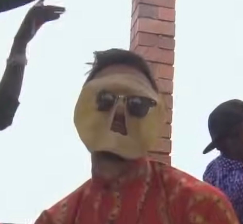 Cropped screenshot from the linked YouTube video. Mamdani dances facing the camera with a chapati (flatbread ala roti) on his face with holes for his eyes and mouth. 