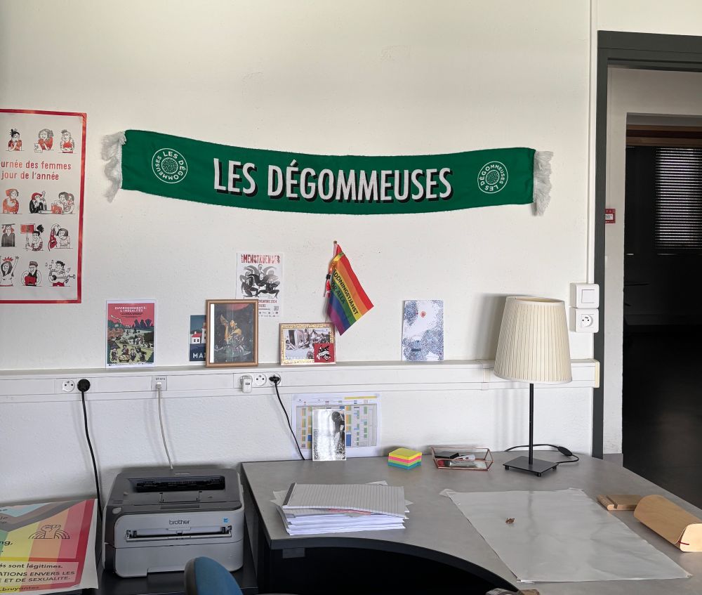 Un bureau professionnel (avec imprimantes, feuilles, post-it, lampe, etc.) dont les murs sont décorés de petites affiches, d'un drapeau arc en ciel et de l'écharpe de foot verte et blanche de l'équipe des Dégommeuses