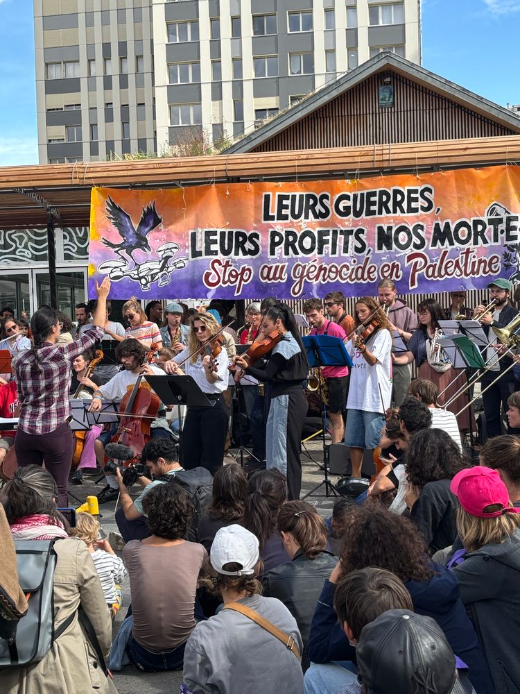 Un orchestre joue devant des gens assis et sous une banderole orange, rose, violet « leurs guerres, leurs profits, nos mortes. Stop au genocide en Palestine »