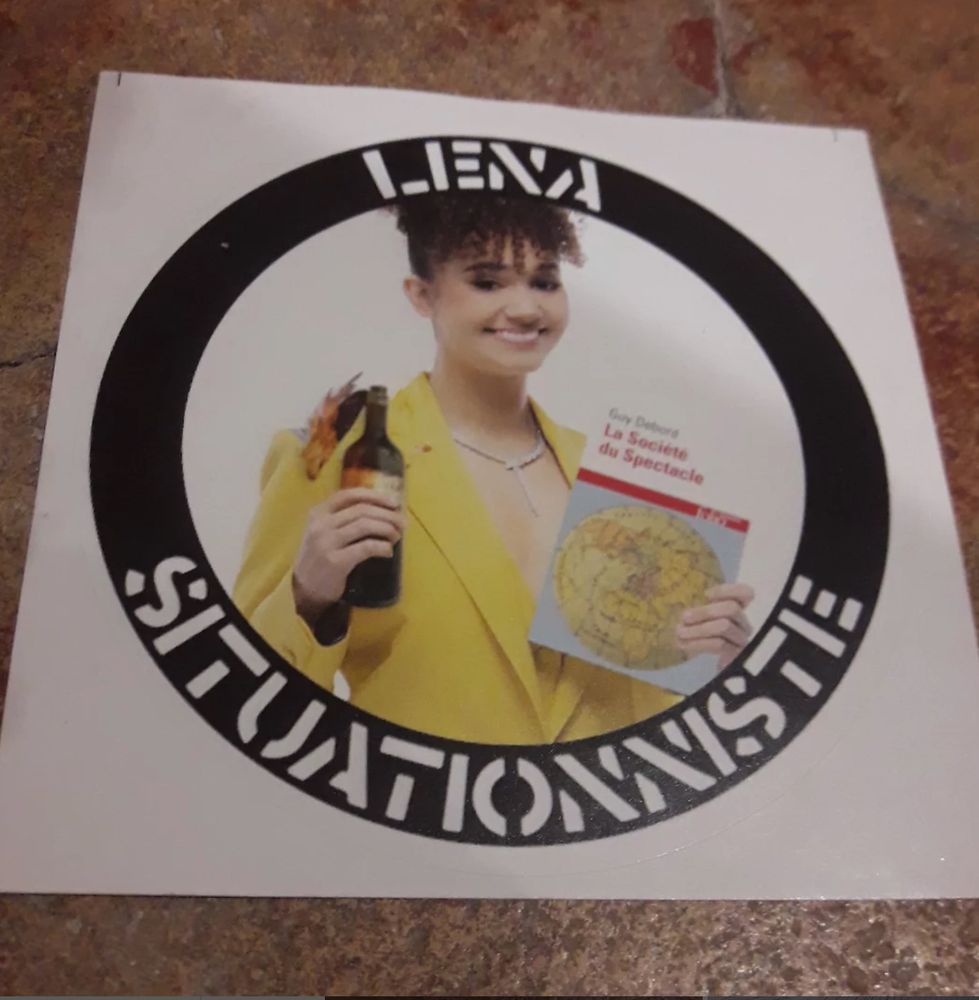 Une affiche représentant la youtubeuse Léna Situation, rayonnante dans une veste jaune et portant un collier en diamant, tenant dans sa main gauche l'ouvrage de Guy Debord "La société du spectacle" et dans la main droite un cocktail molotov. La légende entourant son portrait blanc sur fond noir : "Léna situationniste" 