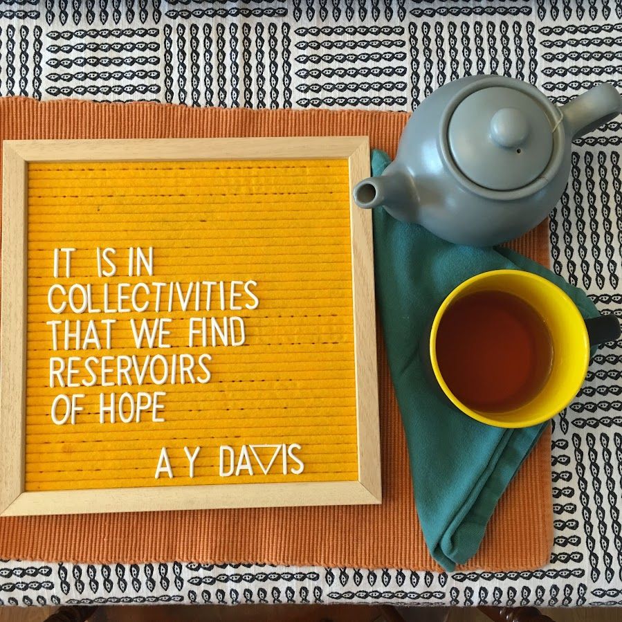 Flay lay-style photo of a blue and yellow teacup, small grey teapot set atop a teal cloth napkin on an orange placemat. They are placed to the right of a bright yellow felt letterboard with an Angela Davis quote that reads, "It is in collectivities that we find reservoirs of hope."