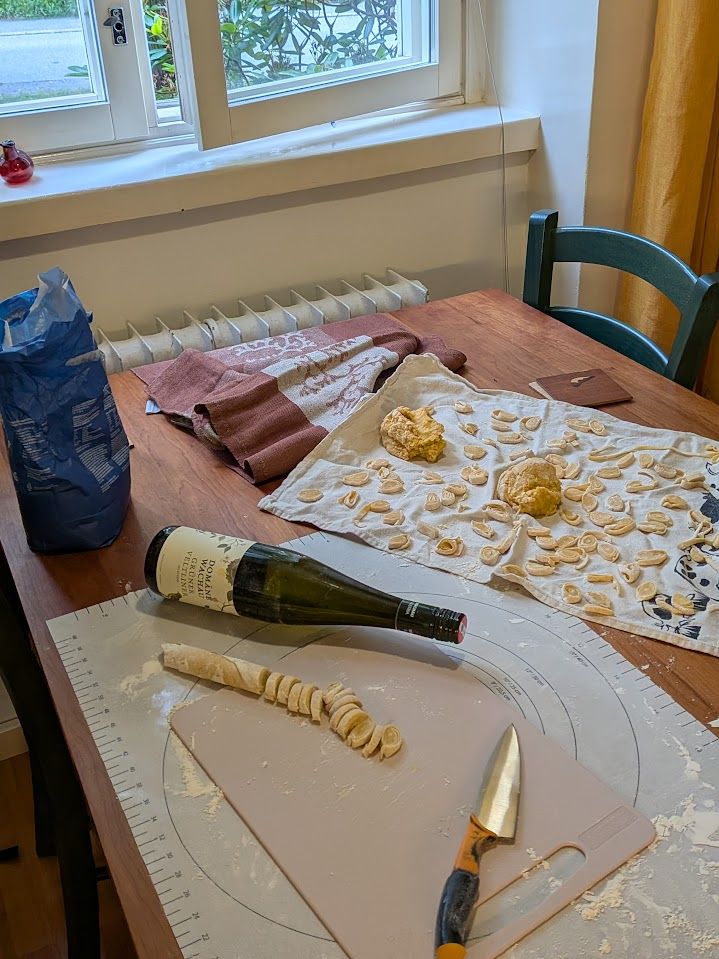 A picture of pasta being made by first flattening the dough with a wine bottle, then rolling the dough, then cutting the rolled dough into strips.