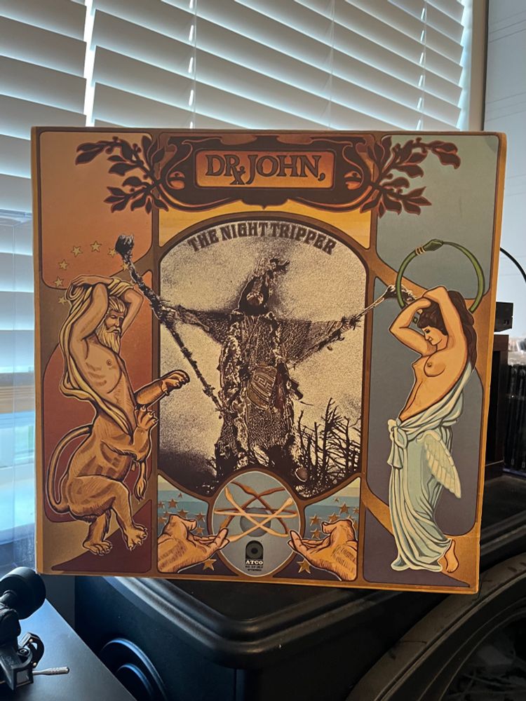 Album cover of "The Sun, Moon & Herbs" by Dr. John, featuring a surreal illustration by John Millerburg of a half-man/half-lion on the left, a topless woman with wings on her robe on the right, and Dr. John as the Night Tripper in the middle.