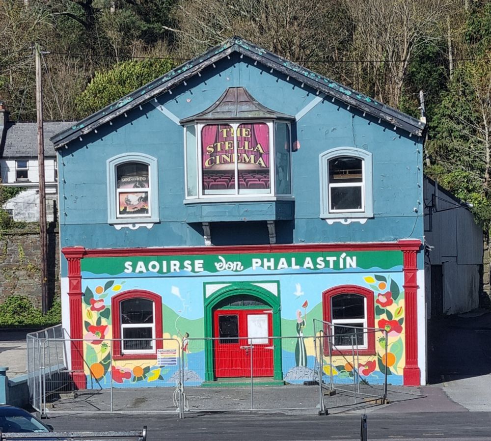 The gable end of a former cinema, painted with a mural covering the whole ground floor, with green, red, flowers, and "saoirse don phalastín" (free palestine). A sunny day. The old sign reads "the stella cinema"