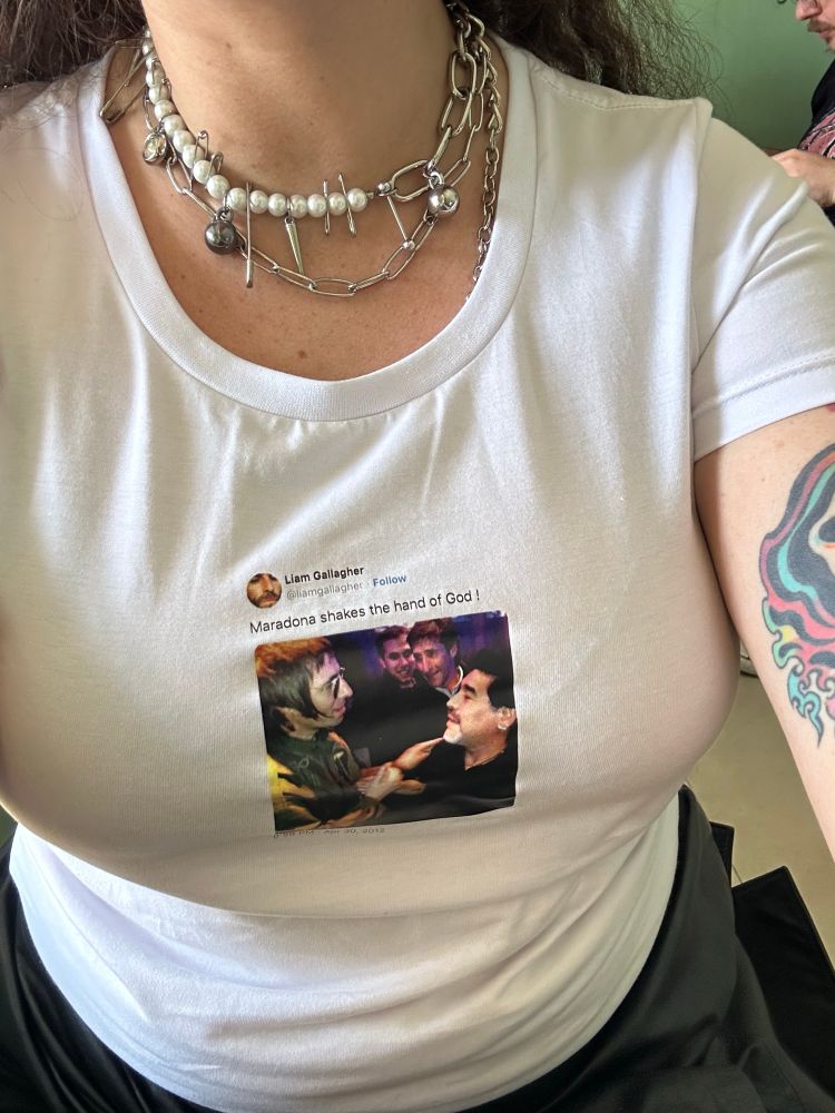 Foto do meu torso. Uso uma camisa branca com a estampa de um tweet de Liam Gallagher dizendo: Maradona shakes the hand of God ! Seguido por uma imagem dos dois.
