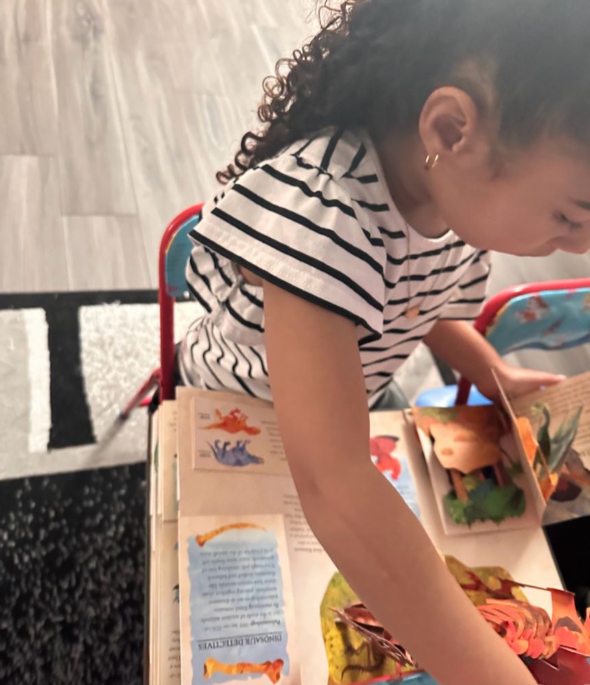 A young child with curly hair and a striped shirt is sitting at a small table, exploring Robert Sabuda’s Encyclopedia Prehistorica: Dinosaurs, a colorful pop-up book featuring detailed dinosaur illustrations and intricate pop-up designs. The child is focused on interacting with the book’s dynamic pages, and the setting includes a tiled floor and part of a rug in the background.
