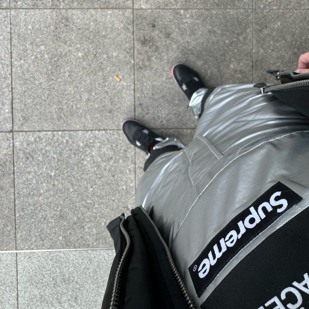 Standing up POV down an open jacket and a shiny silver bib with North Face and Supreme brand labels to black and grey Nike Air Jordan 4.