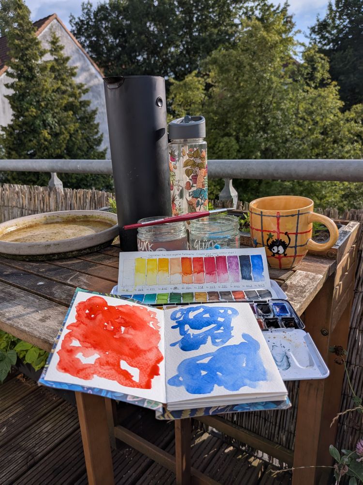 Zu sehen ist ein Balkontisch. Darauf stehen eine Teekanne, eine Wasserflasche, eine Teetasse, Gläser mit Pinselwasser, ein Kasten Aquarellfarben, ein Skizzenbuch mit Farbblobs in Rot und Blau und zuletzt ein Pinsel.
