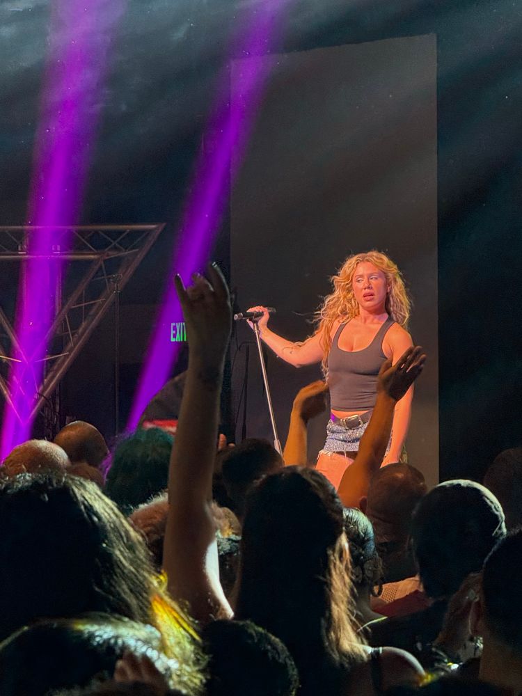 Slayyyter on stage holding the microphone stand with purple lights behind her