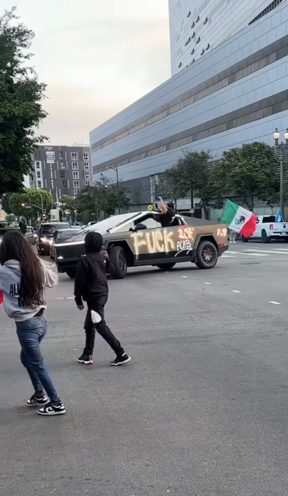 A cybertruck in La with “FUCK ICE. ACAB” spray painted, a man hanging out the window, and a Mexican flag flying from the trunk 