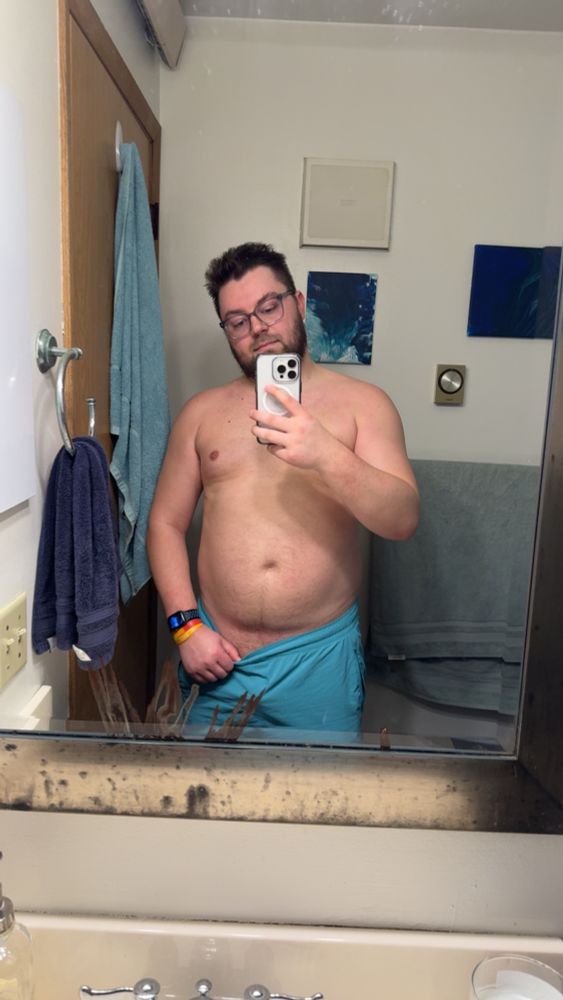 Me standing sweaty & shirtless in the mirror holing my white phone wearing glasses and teal shorts 