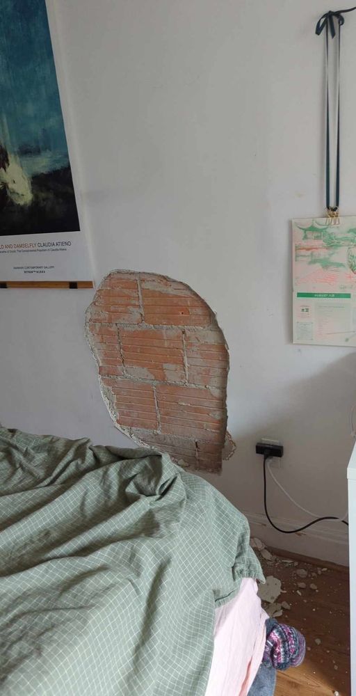 A photo showing a rumpled corner of a bed, the sliver of a poster for podcast "Within the Wires", a 2023 MAU calendar by an artist's collective in Vancouver, and a huge, gaping hole in the wall with several stacked bricks behind it.
