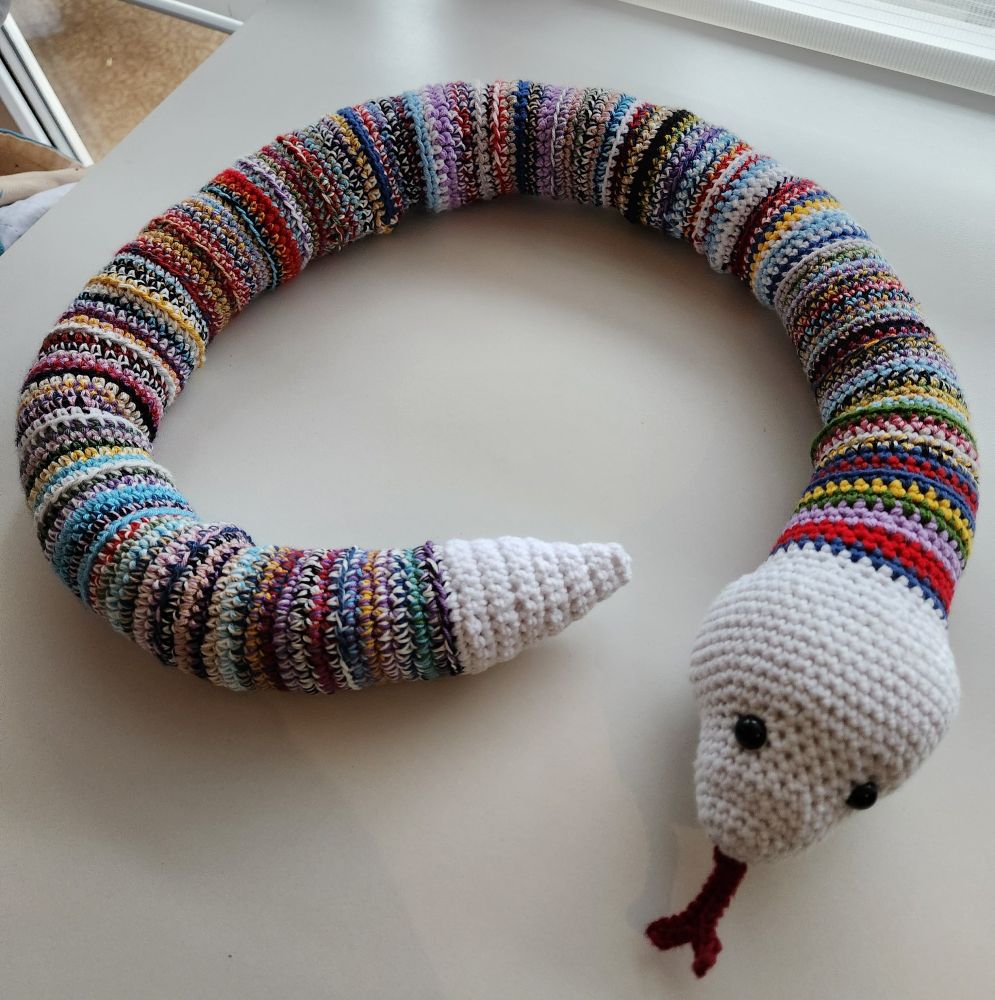 colorfully striped crochet snake with a white head, tongue sticking out, and white tail end laying on a white underground.