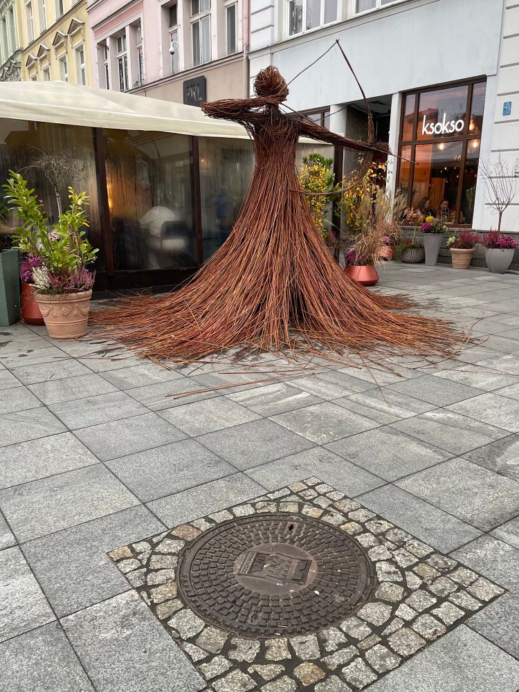 A larger-than-life straw sculpture of a female archer stands on a town square with an outdoor cafe and some pastel-coloured tenement blocks behind it and a manhole cover with a cobblestone surround in front. 