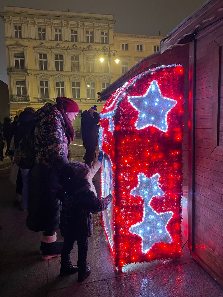 A side view of the illuminated Christmas post box in Bydgoszcz, covered in red and white lights with glowing star decorations. A young child, bundled up in winter clothes, reaches towards the Santa illustration while an adult stands close by. Historic buildings and soft street lighting create a cosy festive atmosphere in the background.