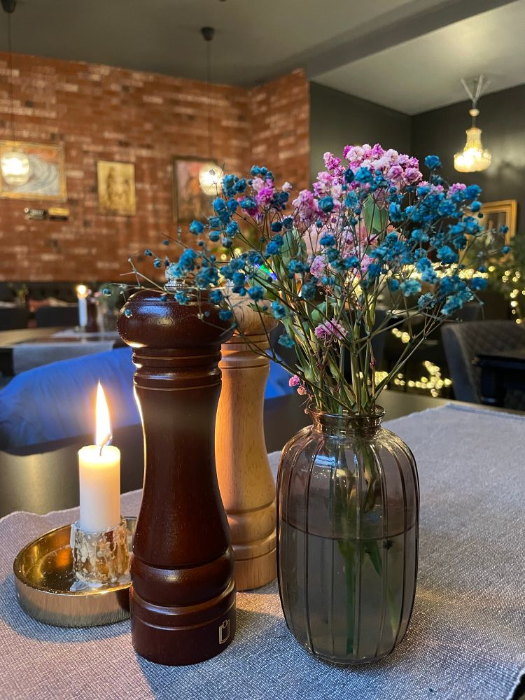An attractive table decoration of a vase with dried flowers, a salt, and pepper, shaker and a lit candle. In the background is a brick design wall with paintings of horses.  