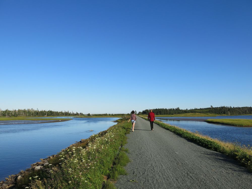 Two people walking away from the camera on a flat gravelled trail with bright blue sky and blue water around them.