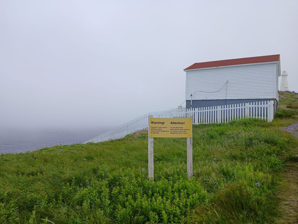 A small white building with a red roof on a hill surrounded by fog, a yellow sign warns that the fog alarm may sound at any time.