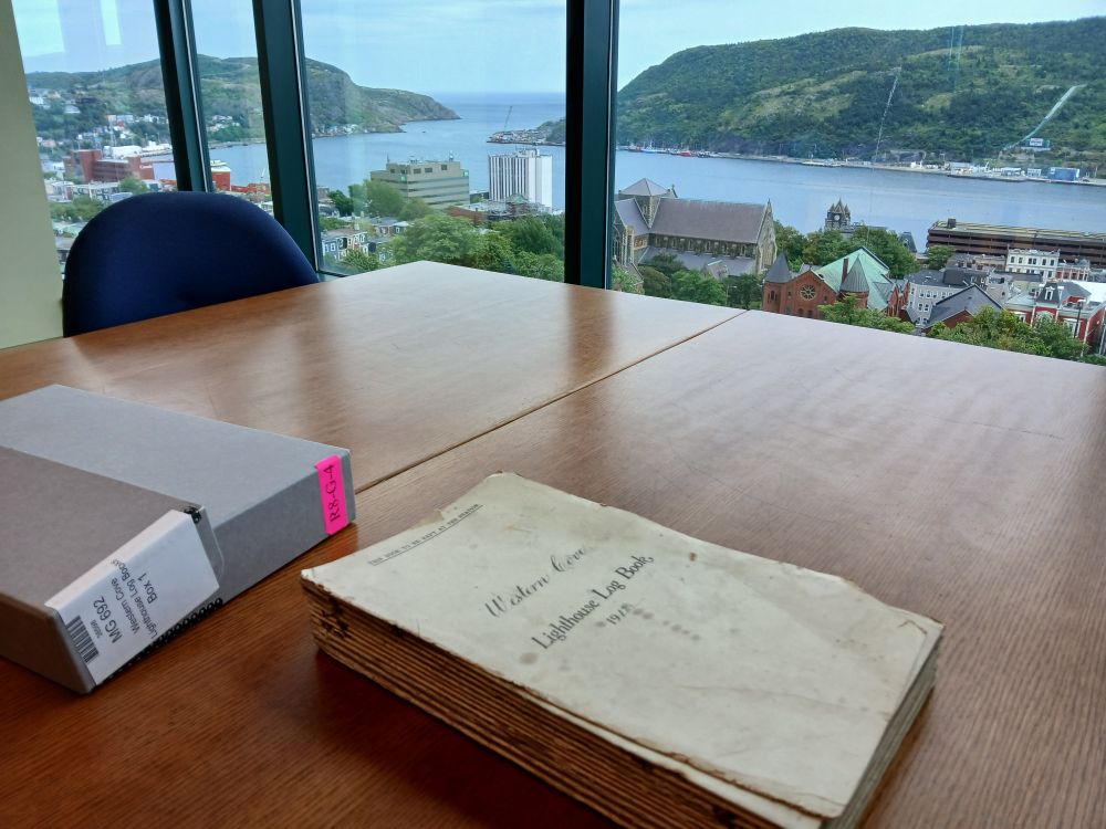 A pile of yellowed paper on a wooden table beside a big window looking out over St. John's harbour.