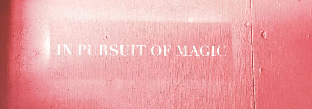 A smooth wooden board painted bright red with white-lettered text reading "IN PURSUIT OF MAGIC".