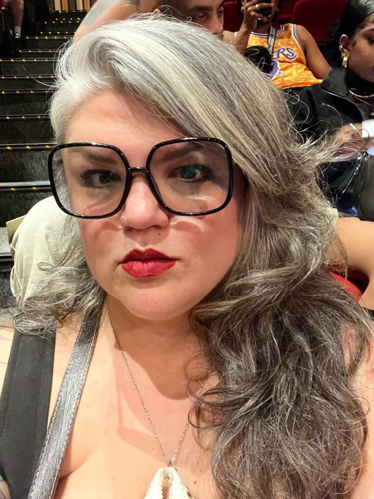 Selfie da minha pessoa, mulher branca de cabelo comprido ondulado grisalho com óculos quadrado preto e batom vermelho. Não estou fazendo bico de pato, meu beicinho é assim mesmo 