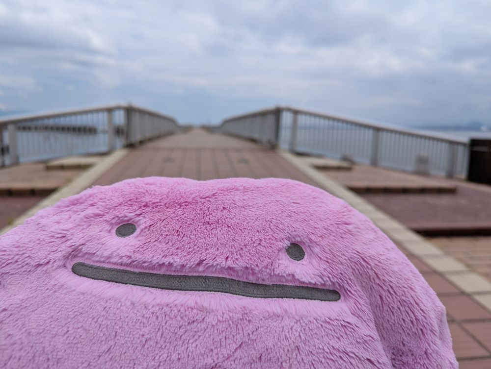 こちらを向いているふわふわメタモンぬいぐるみ。釣り人さんがたくさんいる桟橋の入り口前で撮影。ポケモン世界なら１人は釣り竿をくれるおじさんで他はバトルが始まるトレーナーなんだろうな