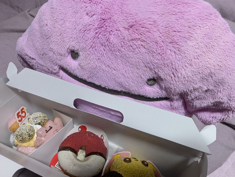 ミスドの箱の中を覗き込むふわふわメタモンぬいぐるみ。メタモンドーナツがこちらを見ている