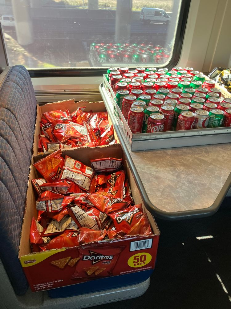 Box of Doritos and other food, and a pallet of soda, sitting on the seats and table of a train carriage 