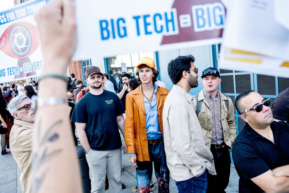 A group of men wait in line for Peter Thiel's lecture, as protesters hold signs criticizing big tech. The man in the center of the frame is a tall thin white man wearing creatively painted jeans, a blue button down with multiple buttons undone, a very large star of david medallion visible beneath his open shirt, an orange leather blazer with large 70s collar, and an orange trucker cap that says "RETARD" in white block letters.