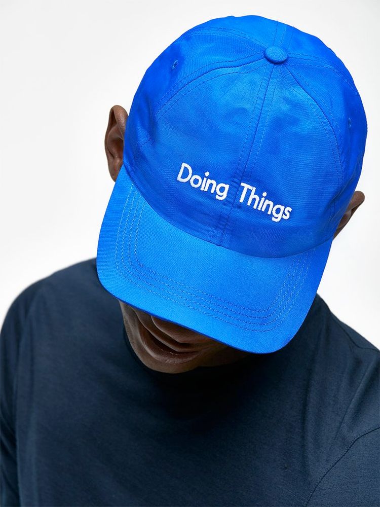 Promotional picture of a bright blue cap with the words Doing Things embroidered on white. The hat is worn by a dark skinned model whose eyes are obscured by the hat's brim. 