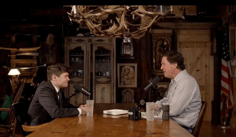 Nick Fuentes talking to Tucker Carlson on his show, facing each other across a wooden table. A chandelier made of antlers hangs above them. In the background, a bunch of creepy woody props line the walls, like oars, tools, an American flag, a hutch with mugs in it, a portrait of a dog, etc.