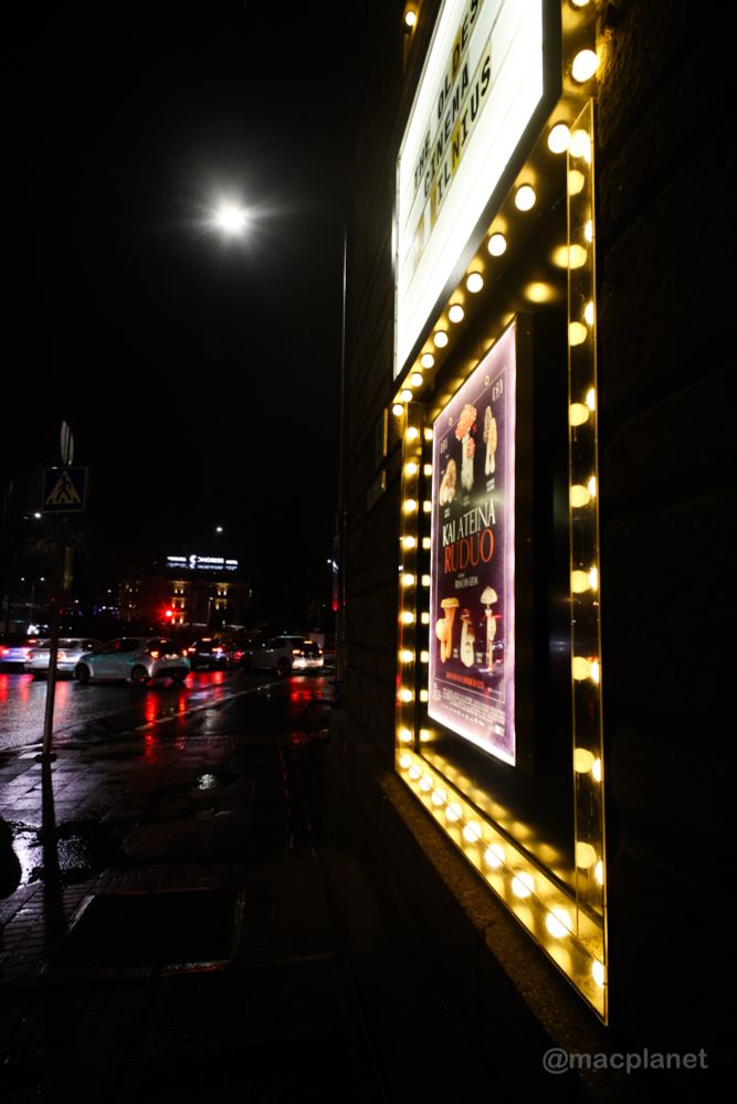 Blick auf die Fassade des Kinos, im Vordergrund ein von Lichtern umrahmtes Filmplakat, im Hintergrund eine regennasse Straße. 