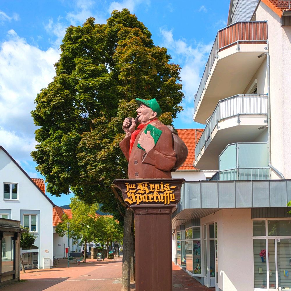 Metallene Wegweiser-Säule in einer Fußgängerzone, oben mit einem hölzernen Wandrersmann mit Pfeife im Mund, grüner Kappe auf dem Kopf und grünem Buch in der Hand mit der Aufschrift "Sparbuch". Darunter in gebrochener Schrift: "Zur Kreissparkasse".