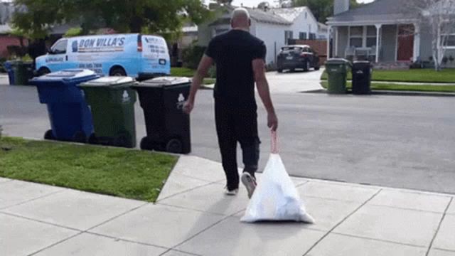 a man is carrying a bag of trash down a sidewalk in front of a don villas van