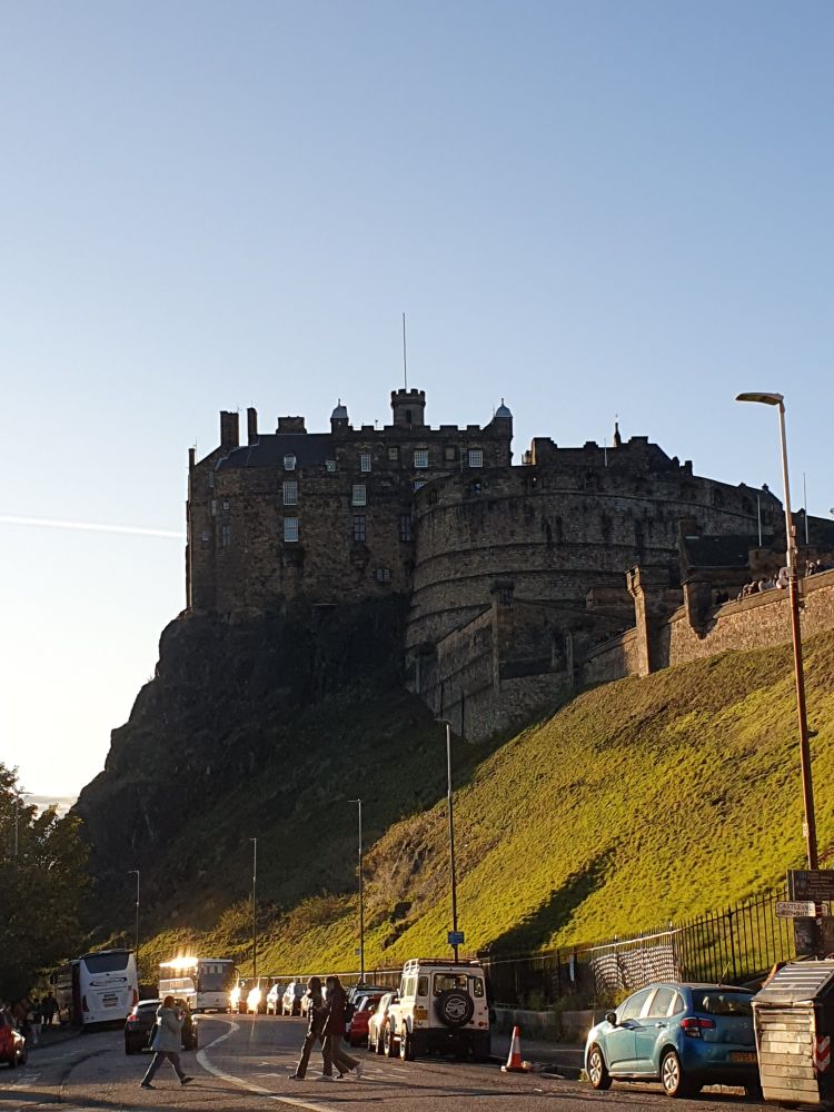 Edinburgh castle against a pale blue sky, with the grassy slope below lit up in the sunlight and the left of the castle rock in shadow.