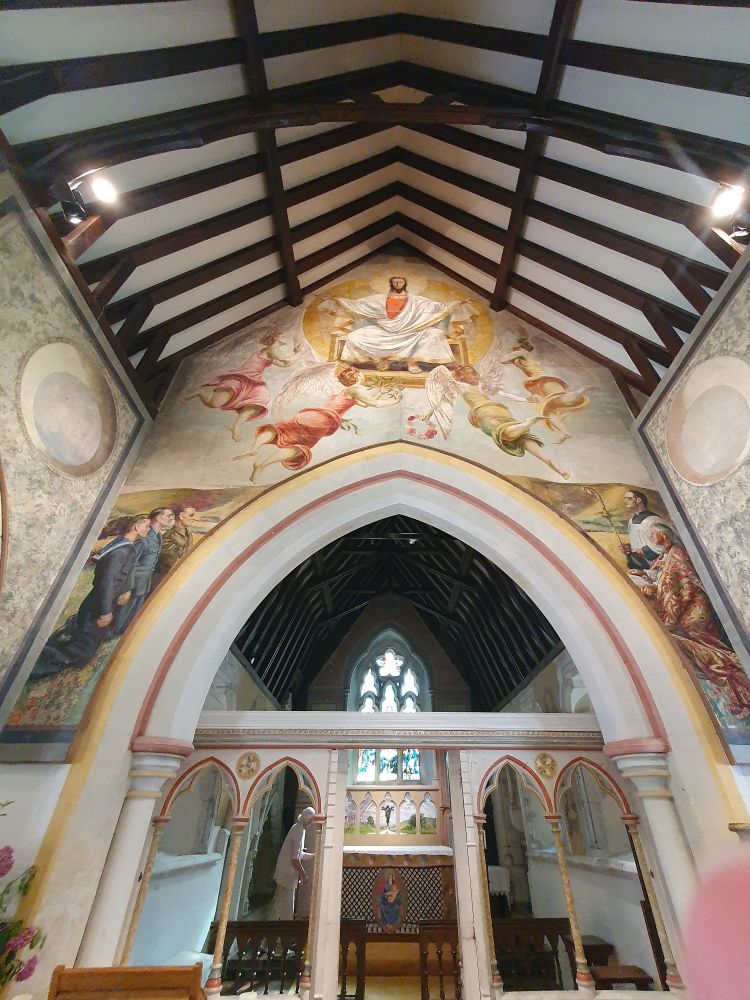 Mural above the arch of the inside of a church, facing the altar, showing Jesus in the centre with three military figures kneeling on the left and church figures on the right. Dark wooden beams supporting the white-painted ceiling.