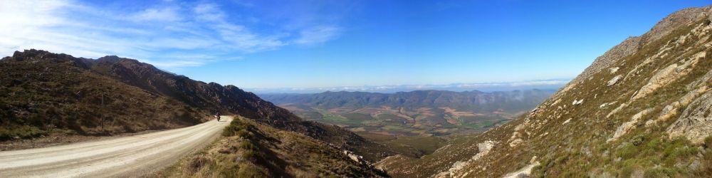 A panorama of the Swartberg Pass near Prince Albert, South Africa, showcasing a winding dirt road flanked by rugged mountain slopes and a vast valley stretching into the horizon under a bright blue sky