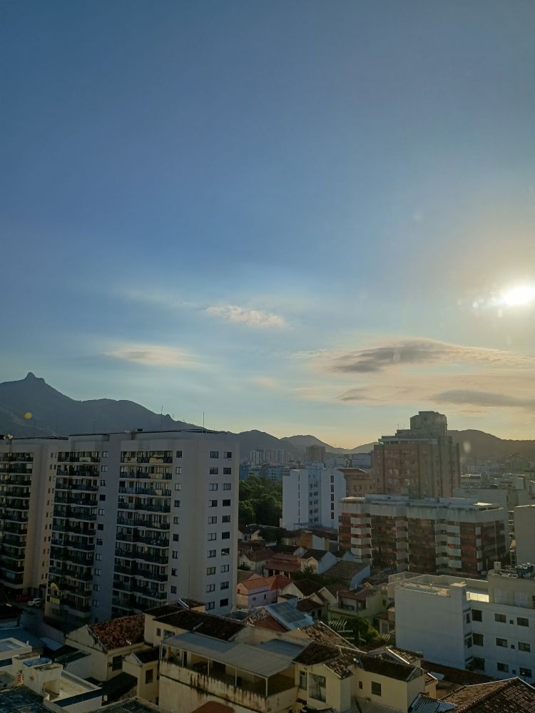 Foto tirada do alto, mostra vários prédios e casas das redondezas embaixo de um céu azulado iluminado pelo sol do fim do dia.