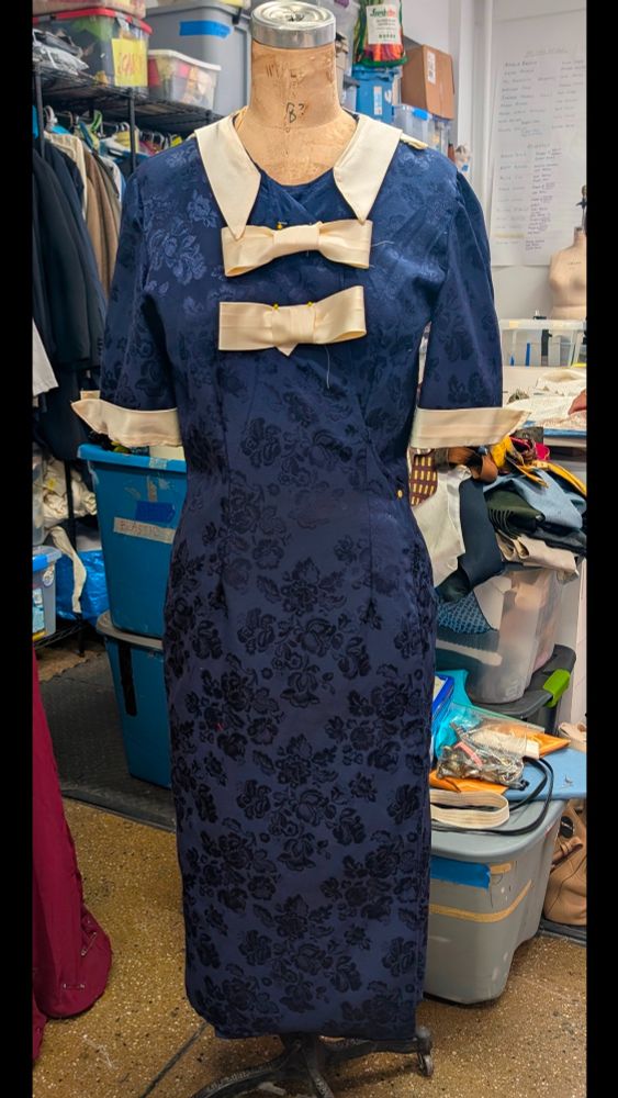 A navy blue dress with a floral print and cream ribbons for bows and a collar. Dress is on a dress form in a costume workshop.