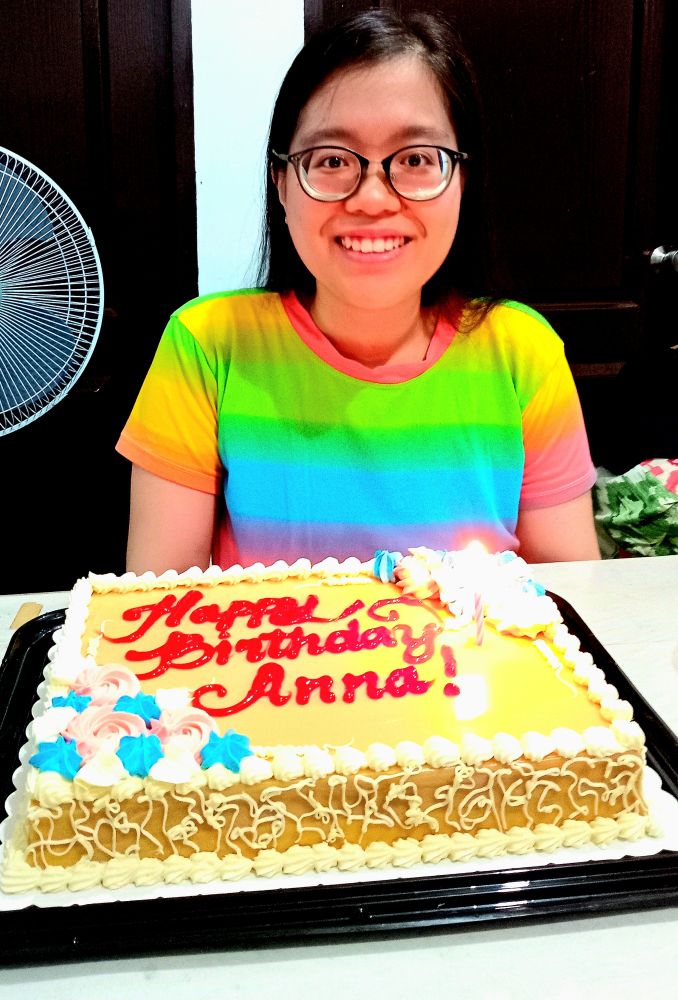 Anna wearing a rainbow shirt. She has a birthday cake in front of her that reads "Happy Birthday Anna!"