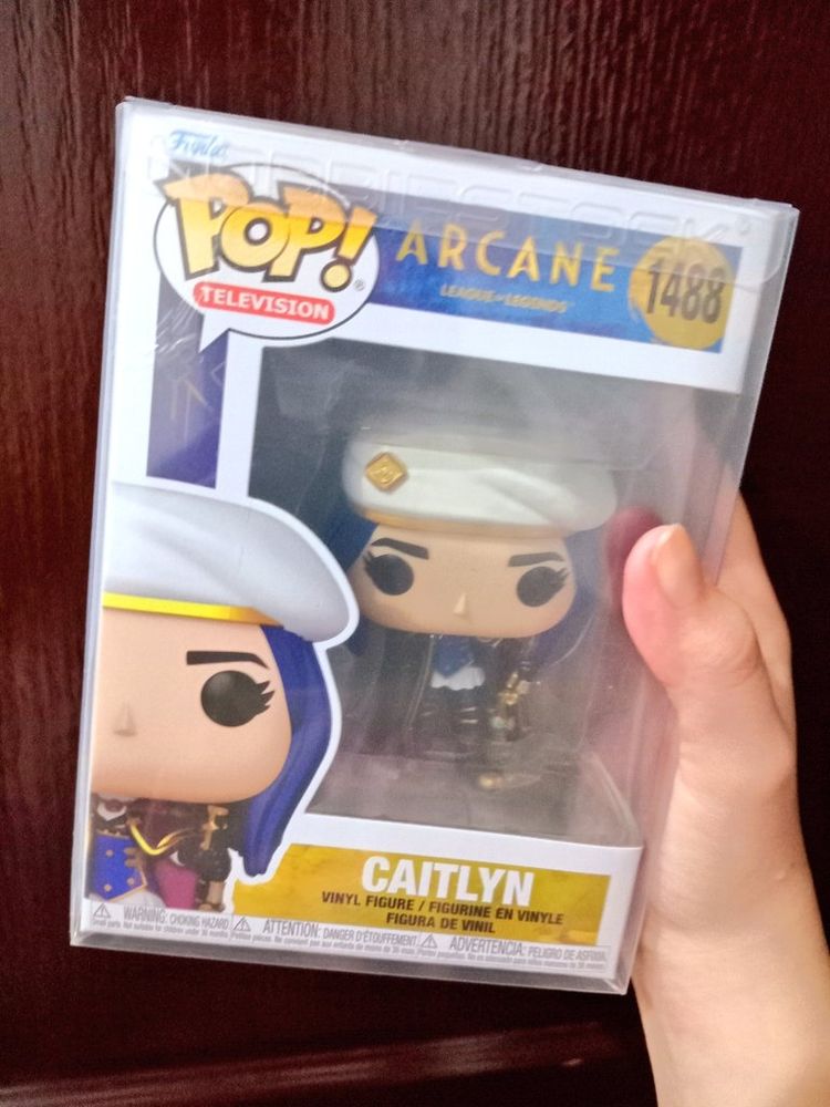 Anna's hand holding the Caitlyn Kiramman Funko Pop against a wooden background. It is still in the box and it has a plastic protector.