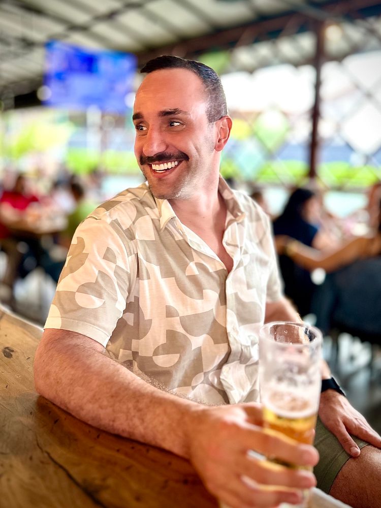 Homem branco sentado em uma mesa de bar. Ele olha para o lado e sorri. Ele usa uma camisa branca com bege, tem bigode e segura um copo de cerveja com a mão direita. 