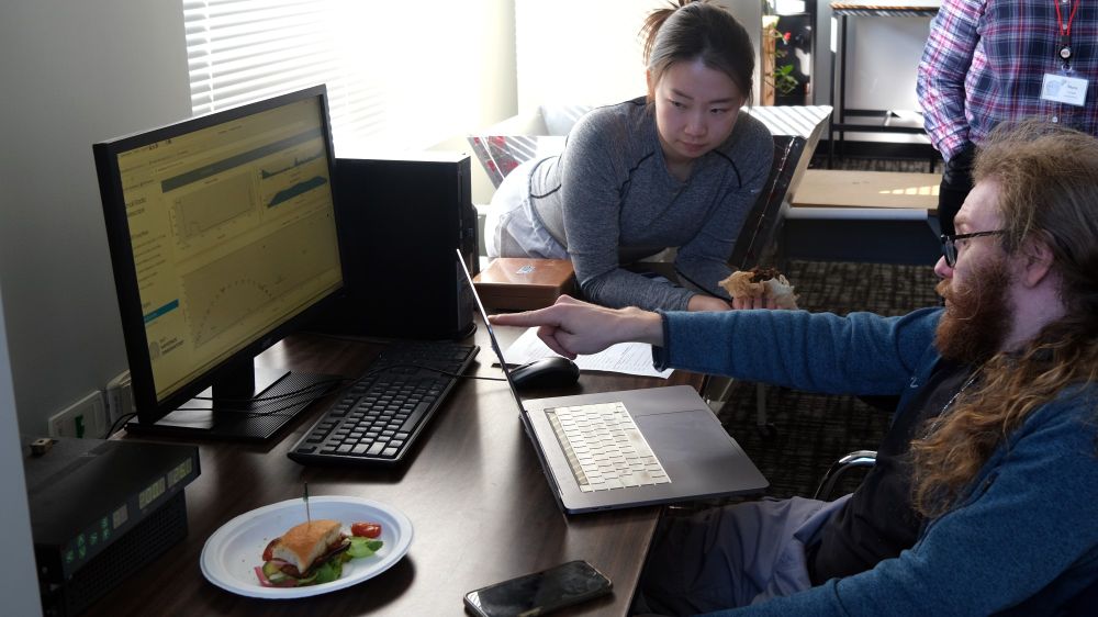 two people looking at a computer monitor with a laptop and a sandwich on the table; one person is pointing at the screen