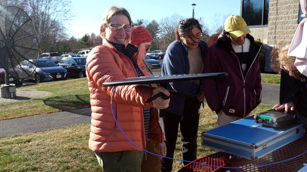 a woman standing next to three people outside, holding a flat black antenna pointed at the camera