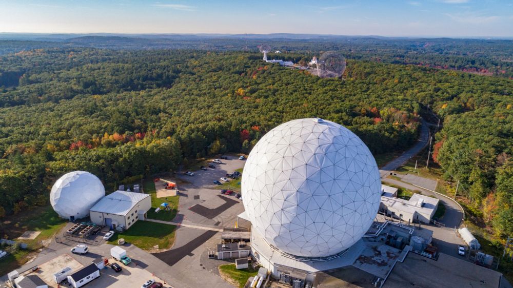 A large white dome-shaped radio telescope enclosure surrounded by green trees; a smaller domed enclosure is off to the left and there are several outbuildings