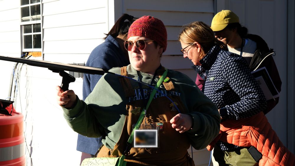 a person in a red hat holding an antenna pointed to the left of the viewer, with three others in the background, standing outside