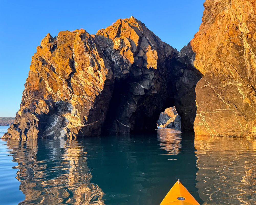 This is a photo of a yellow sea kayak nearing a natural arch off the coast of Pembrokeshire, Wales at 9pm on a fabulous blue sky evening in July 2025. The rocks - which are reflected in the calm water of the sea - are a deep copper colour and the arch provides a window on the cliffs behind.