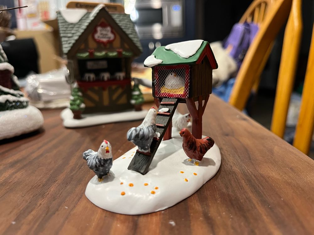 a department 56 chicken coop. there is a chicken in a raised coop, a chicken on a ramp up to the coop, and 3 chickens on the snow. there is also chicken feed scattered. 